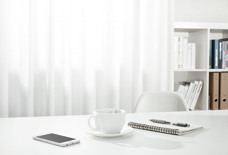 A closeup of a cup of coffee, a notebook and pen, and a smartphone on a white desk, indoors